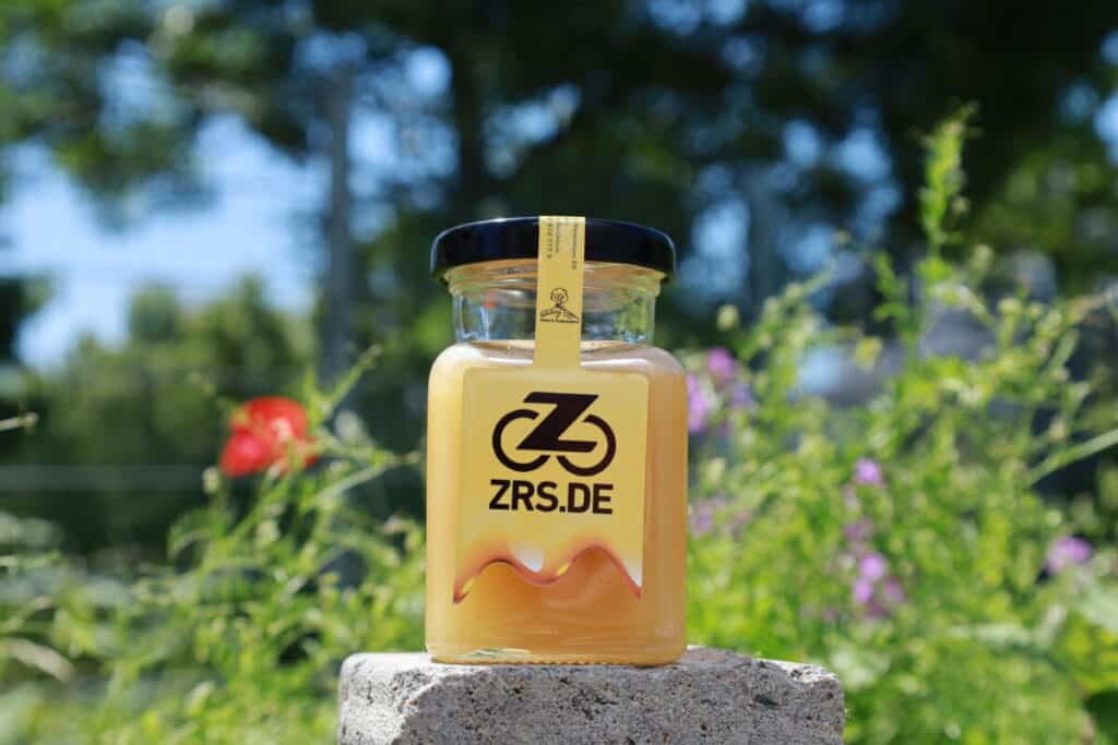 Ein Honigglas mit ZRS-Logo auf einem Stein, dahinter eine Blumenwiese.